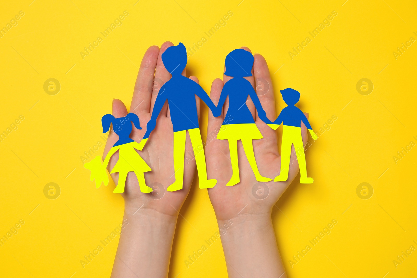 Image of Woman holding paper family figures in colors of Ukrainian flag on yellow background, top view