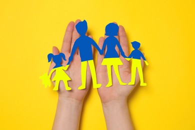 Woman holding paper family figures in colors of Ukrainian flag on yellow background, top view