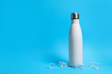 Stylish white thermo bottle with water drops and ice cubes on blue background, space for text