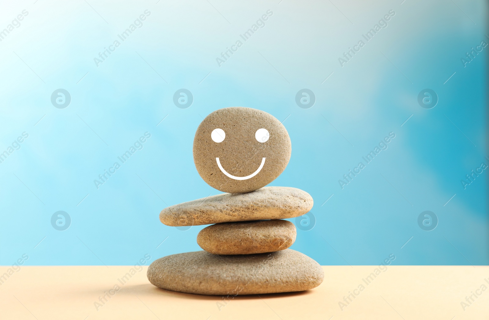 Photo of Stack of stones with drawn happy face on beige table against light blue background. Zen concept