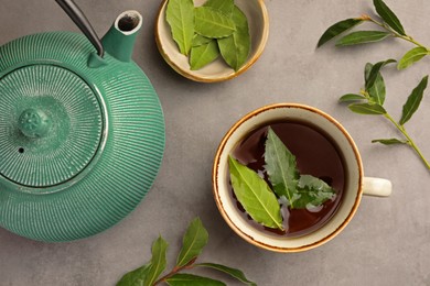 Cup of freshly brewed tea, teapot and bay leaves on grey table, flat lay