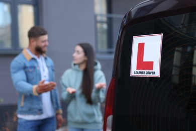 Photo of Learner driver and instructor near car outdoors, selective focus on L-plate. Driving school