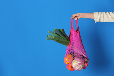 Photo of Woman holding net bag with vegetables on blue background, closeup. Space for text