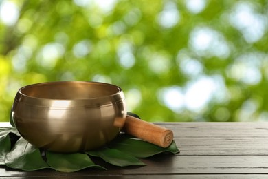 Image of Golden singing bowl, mallet and monstera leaf on wooden table outdoors, space for text