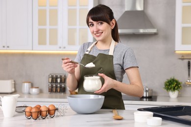 Happy young housewife adding flour into bowl at white marble table in kitchen. Cooking process
