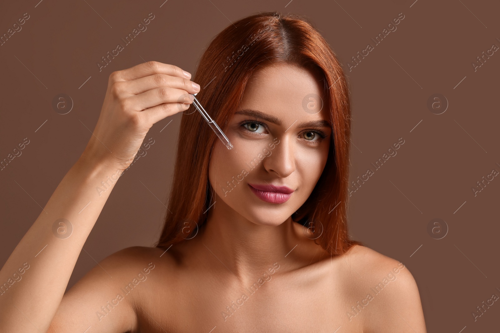 Photo of Beautiful young woman applying cosmetic serum onto her face on brown background