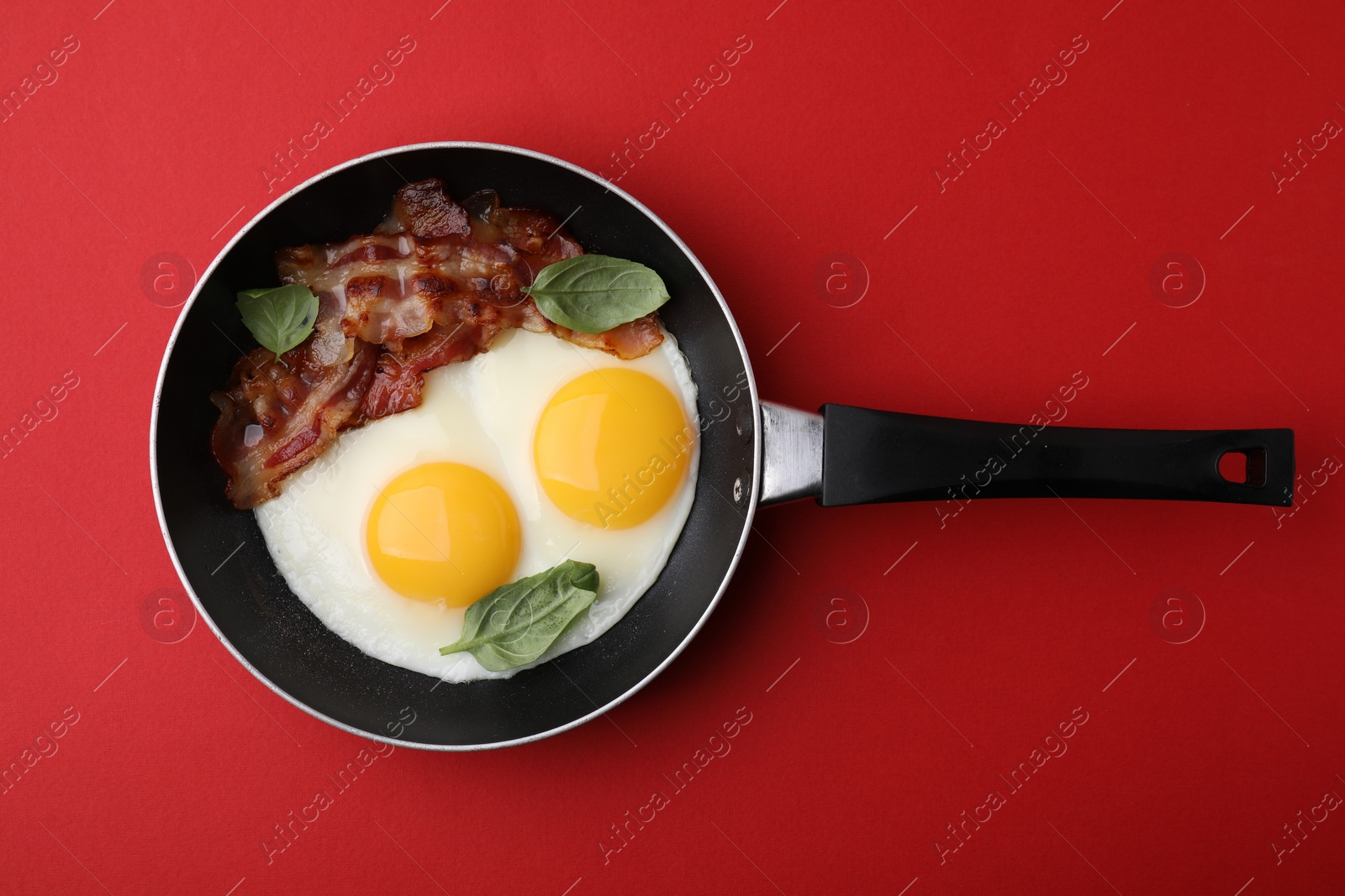 Photo of Fried eggs, bacon and basil in frying pan on red background, top view