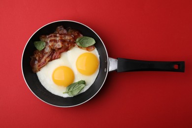 Photo of Fried eggs, bacon and basil in frying pan on red background, top view