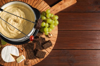 Forks with pieces of grape, bread, fondue pot with melted cheese and snacks on wooden table, top view. Space for text