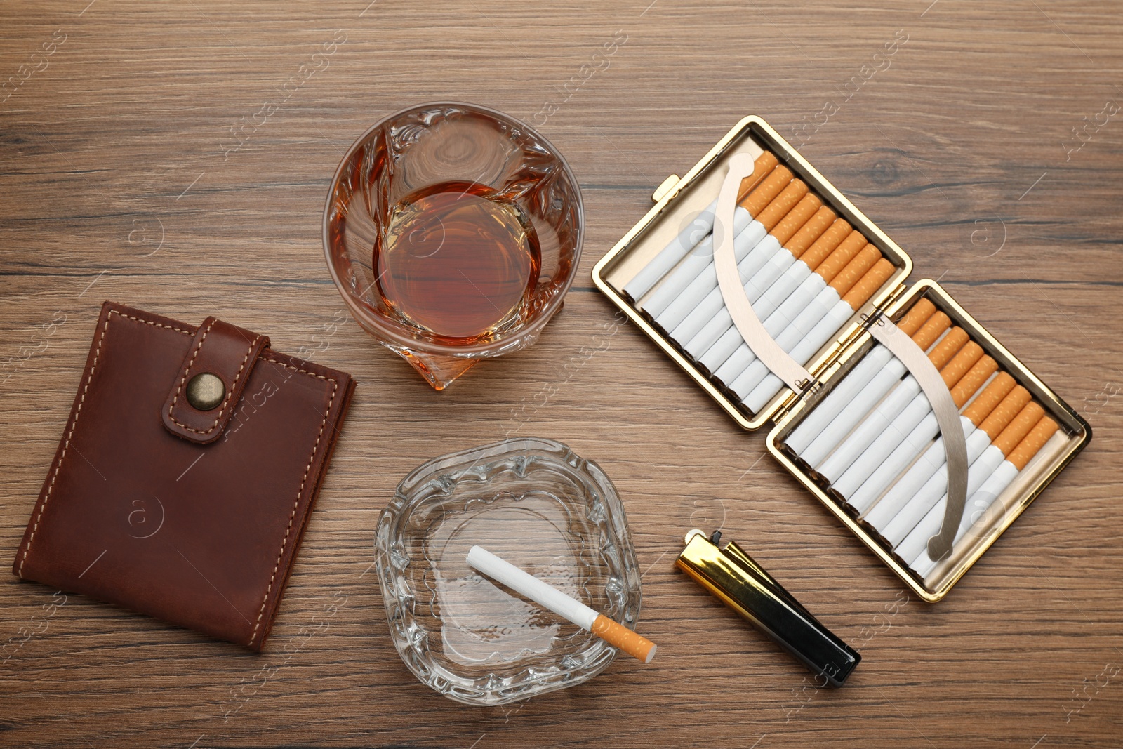 Photo of Open case with tobacco filter cigarettes, lighter, glass ashtray, wallet and alcohol drink on wooden table, flat lay