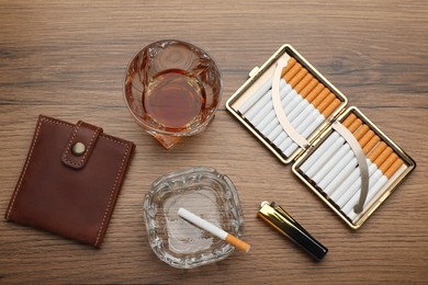 Open case with tobacco filter cigarettes, lighter, glass ashtray, wallet and alcohol drink on wooden table, flat lay