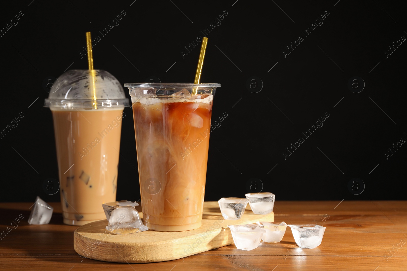 Photo of Refreshing iced coffee with milk in takeaway cups on wooden table against black background, space for text