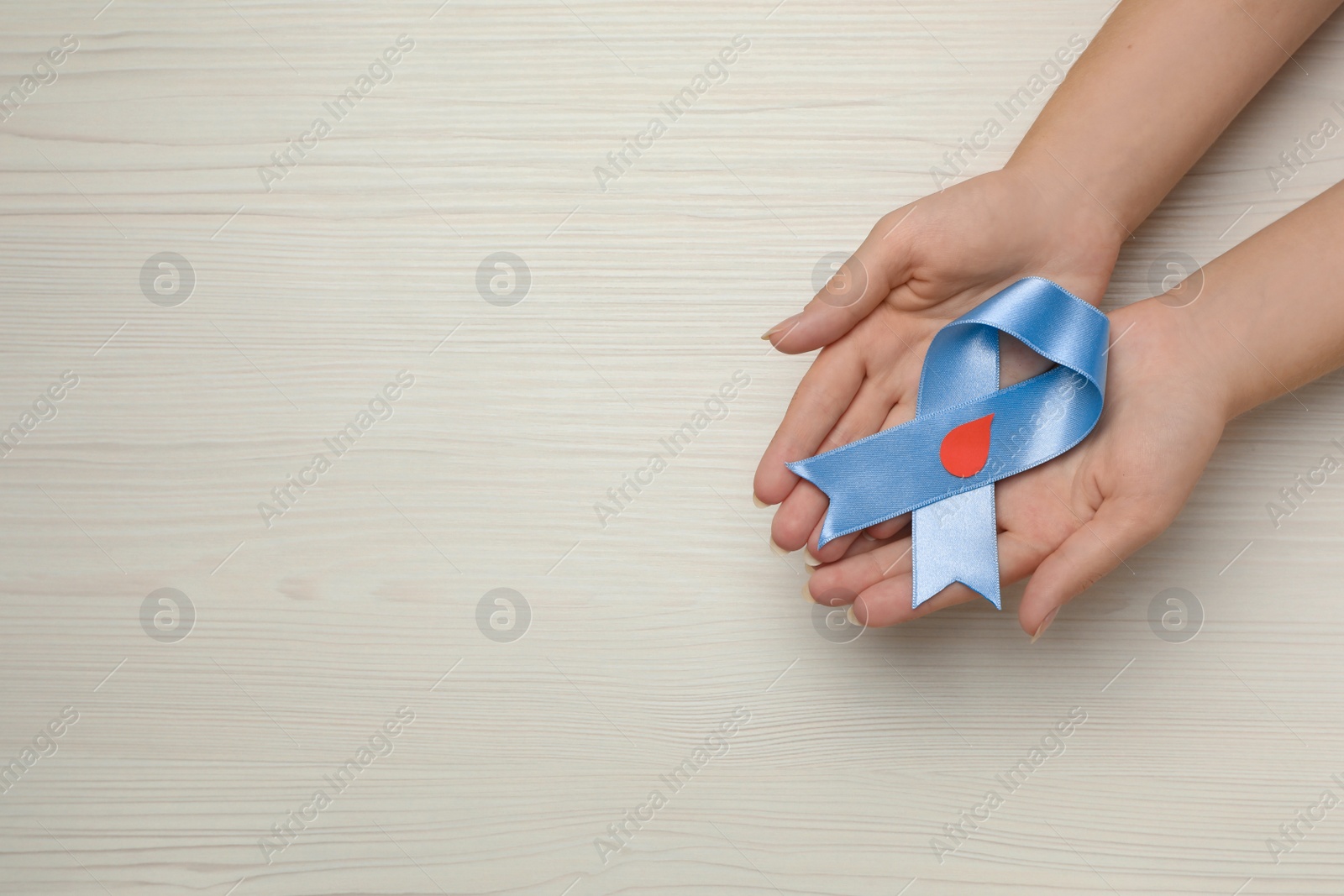 Photo of Woman holding light blue ribbon with paper blood drop at wooden table, top view and space for text. Diabetes awareness