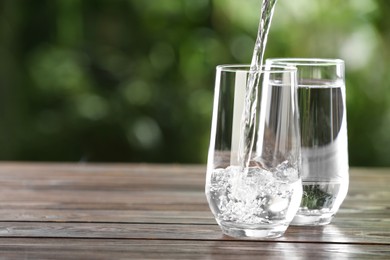 Photo of Pouring water into glass on wooden table outdoors, space for text