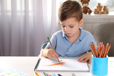 Photo of Cute little boy drawing with pencil at white wooden table in room. Child`s art