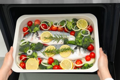 Photo of Woman putting baking dish with raw fish and vegetables into oven, top view