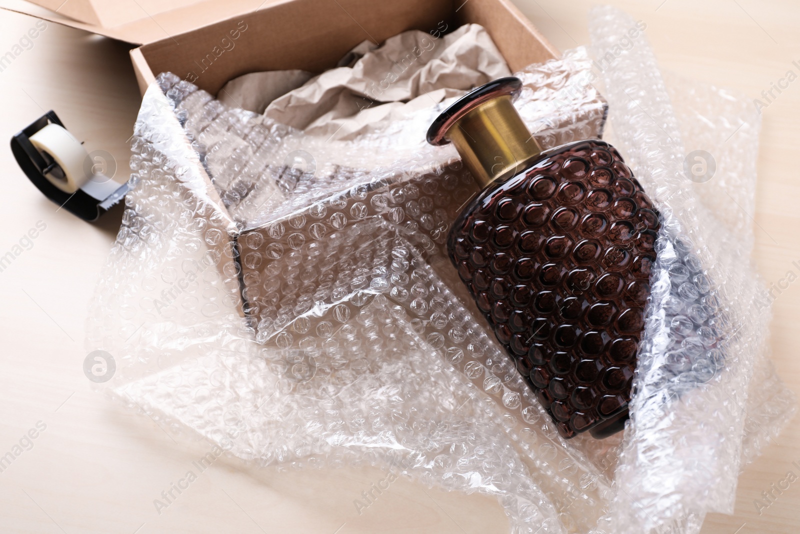 Photo of Beautiful glass vase in bubble wrap near cardboard box with paper and adhesive tape on wooden table