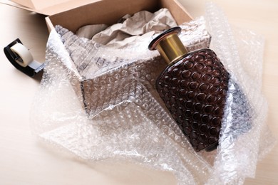 Photo of Beautiful glass vase in bubble wrap near cardboard box with paper and adhesive tape on wooden table