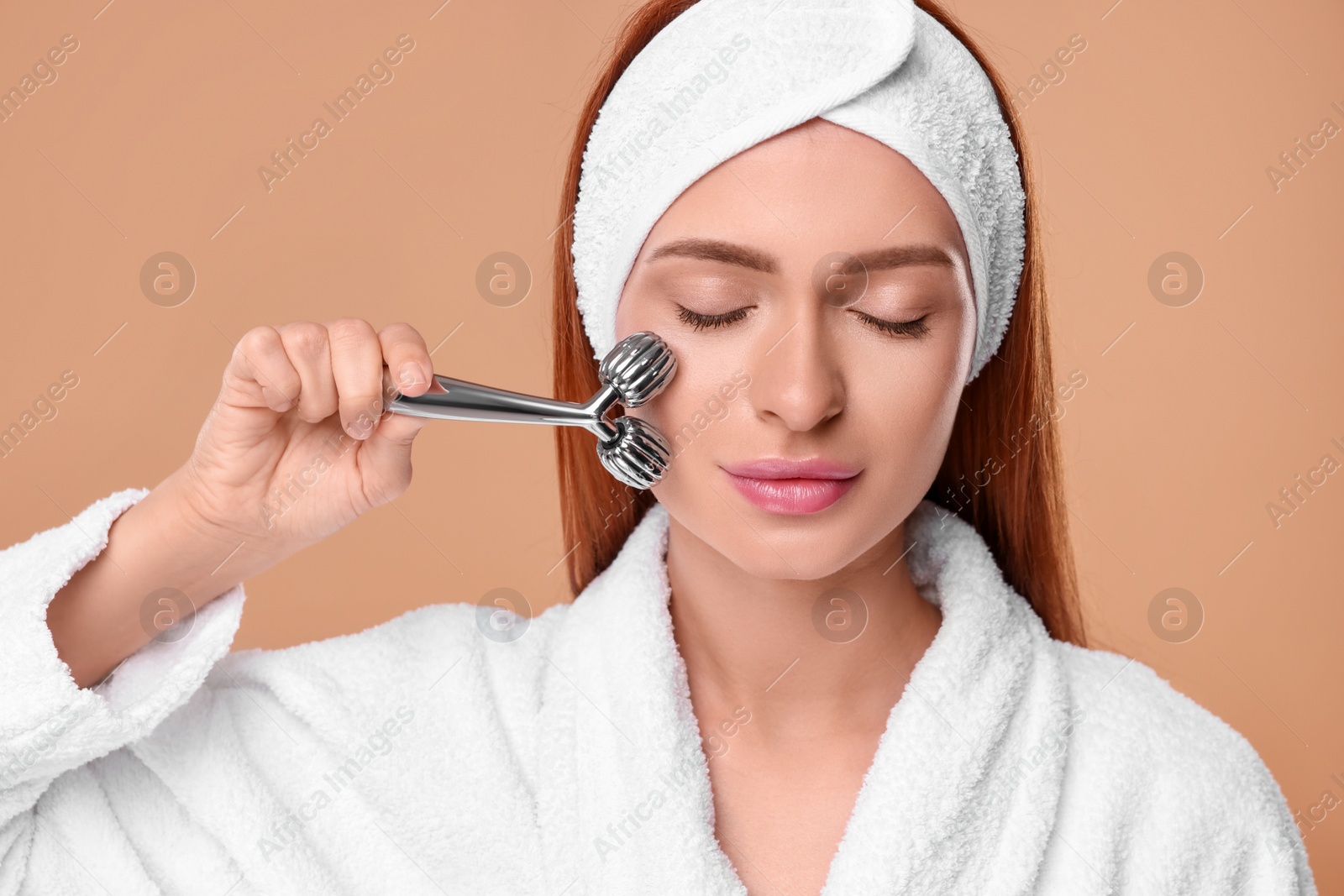 Photo of Young woman massaging her face with metal roller on pale orange background