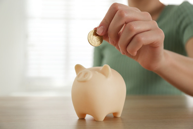 Photo of Woman putting money into piggy bank at wooden table indoors, closeup. Space for text