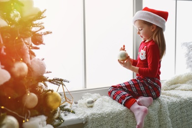 Cute little girl in Santa hat holding Christmas ball on window sill at home