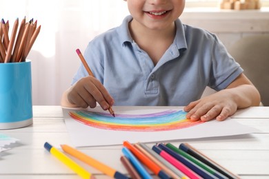 Photo of Little boy drawing rainbow with pencil at white wooden table indoors, closeup. Child`s art