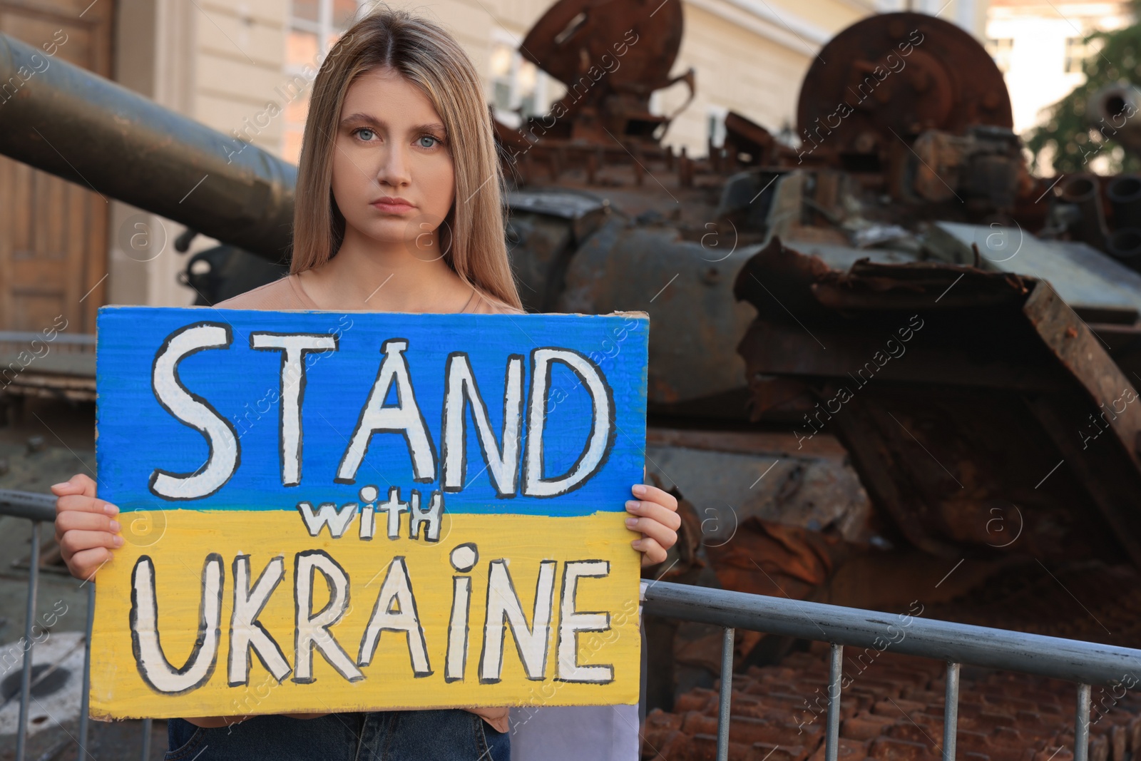 Photo of Sad woman holding poster in colors of national flag with words Stand with Ukraine near broken tank on city street