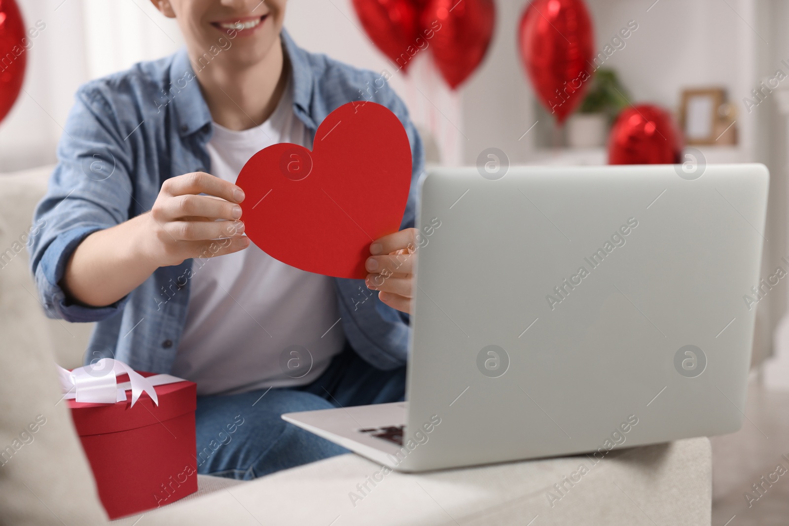 Photo of Valentine's day celebration in long distance relationship. Man holding red paper heart while having video chat with his girlfriend via laptop, closeup