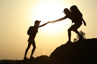 Photo of Silhouettes of man and woman helping each other to climb on hill against sunset