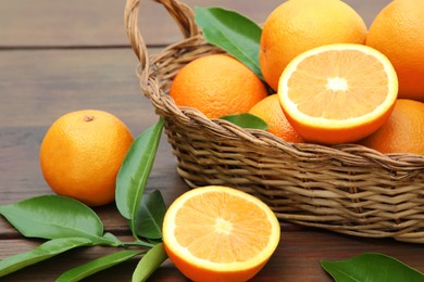 Photo of Wicker basket, ripe juicy oranges and green leaves on wooden table, closeup