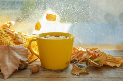 Photo of Cup of hot drink with marshmallows and autumn leaves near window on rainy day. Cozy atmosphere