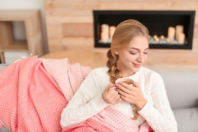 Photo of Beautiful young woman wrapped in plaid sitting on sofa with cup at home. Winter atmosphere