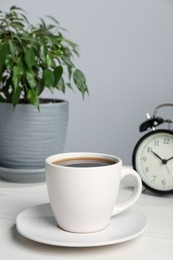 Cup of coffee, houseplant and alarm clock on white wooden table against grey wall