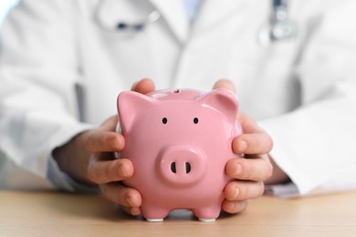Photo of Doctor with piggy bank at wooden table, closeup