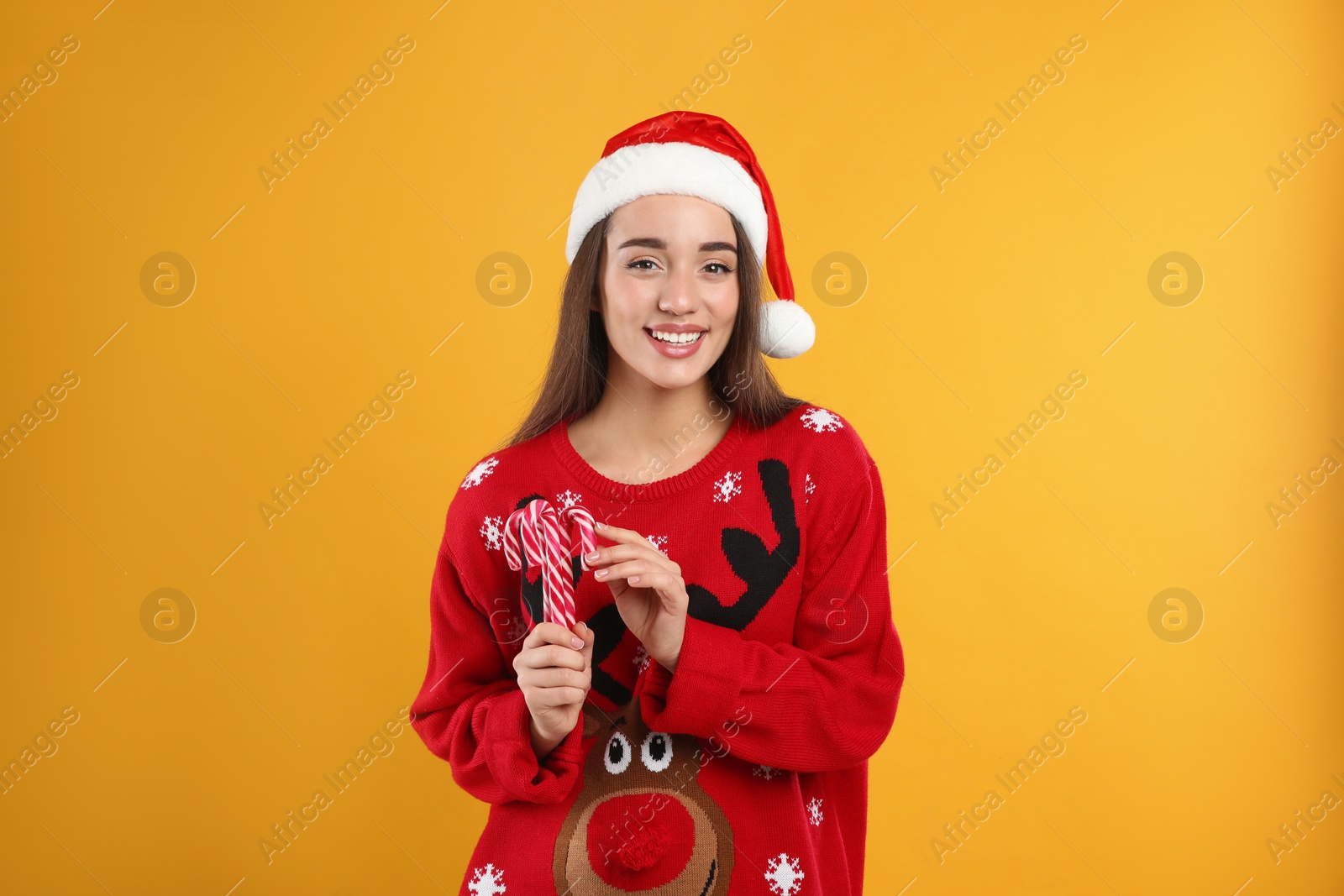 Photo of Young woman in Christmas sweater and Santa hat holding candy canes on yellow background