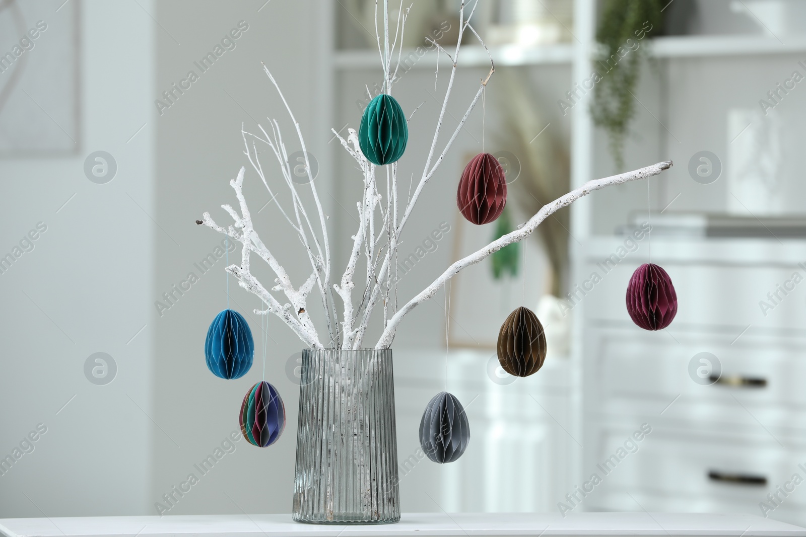 Photo of Branches with paper eggs in vase on white table indoors. Beautiful Easter decor