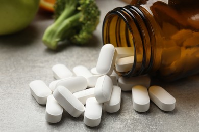 Photo of Dietary supplements. Overturned bottle, pills and broccoli on grey table, closeup