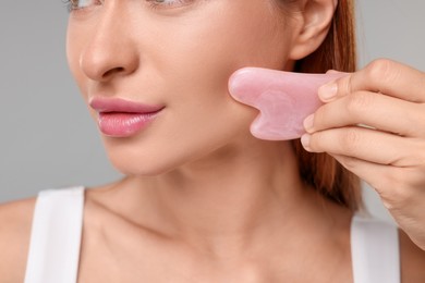 Photo of Young woman massaging her face with rose quartz gua sha tool on grey background, closeup