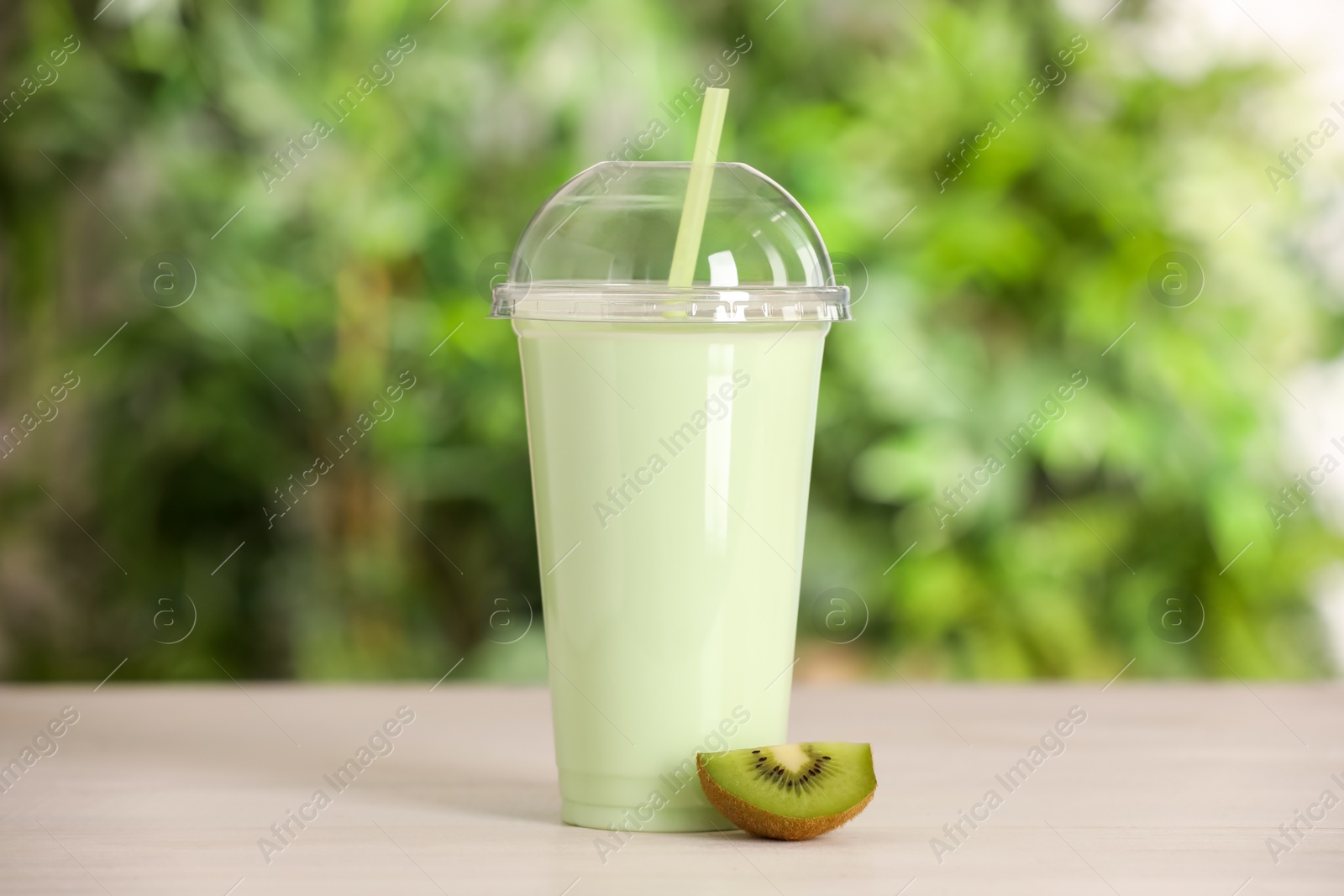 Photo of Plastic cup with tasty smoothie and slice of fresh kiwi on wooden table outdoors