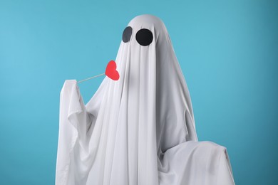 Photo of Cute ghost. Person covered with white sheet holding stick with red heart on light blue background
