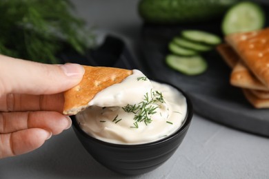 Woman dipping cracker into tasty creamy dill sauce at grey table, closeup