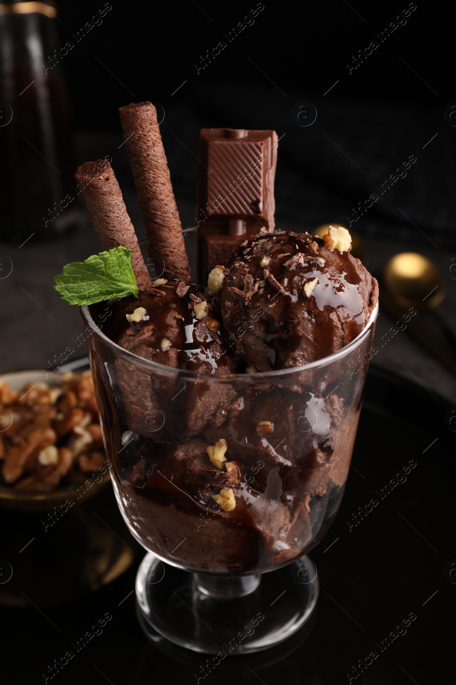 Photo of Tasty chocolate ice cream with sauce, nuts and wafer rolls in glass dessert bowl on table, closeup