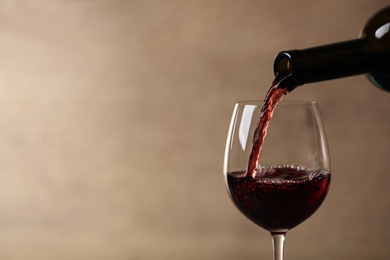 Pouring red wine into glass from bottle against blurred beige background, closeup. Space for text