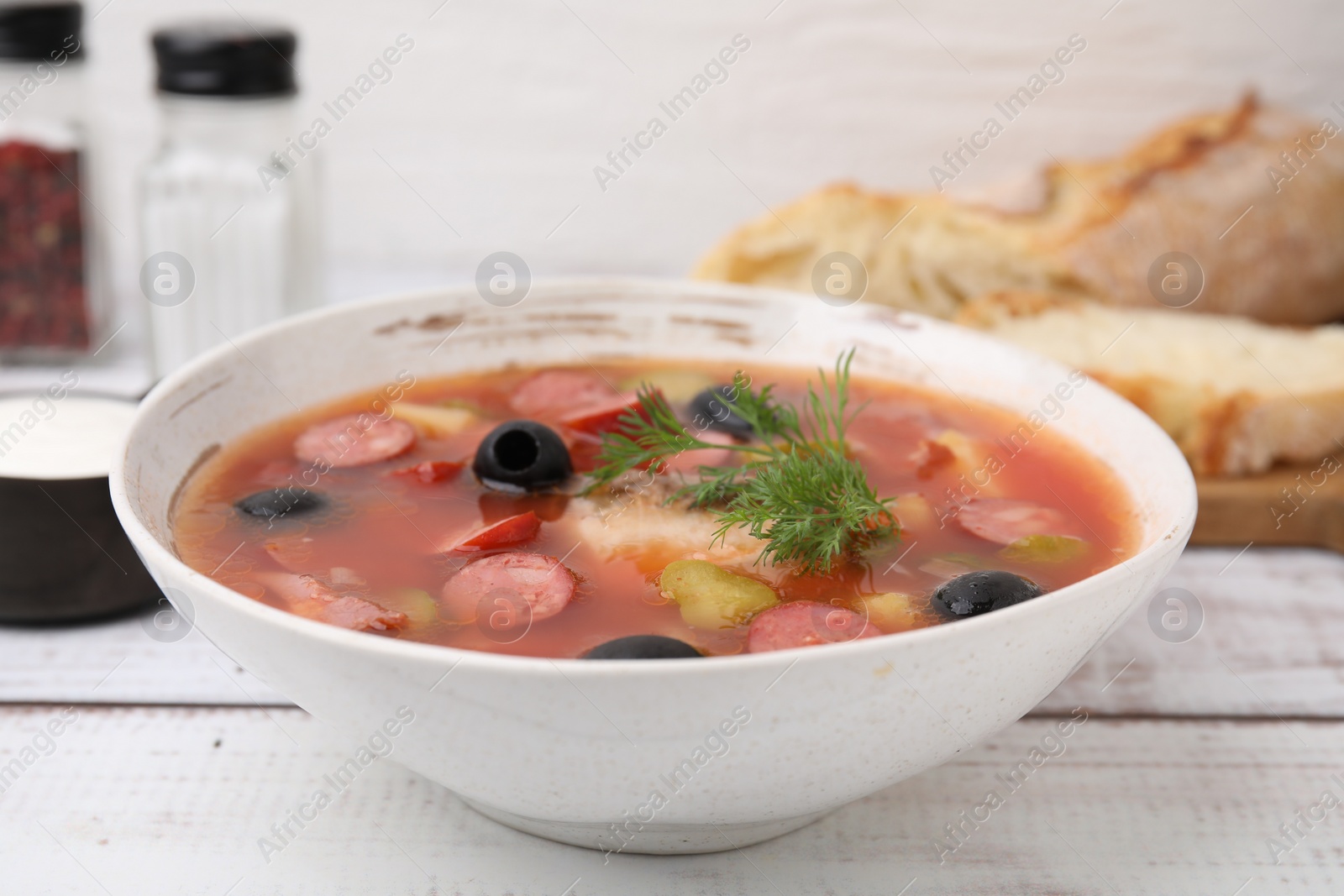 Photo of Meat solyanka soup with thin dry smoked sausages in bowl on white wooden table, closeup