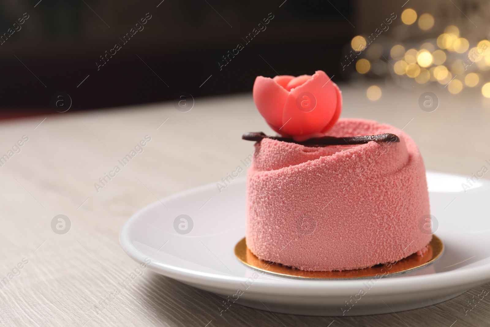 Photo of Delicious mousse cake with floral decor on white wooden table against blurred lights, closeup. Space for text