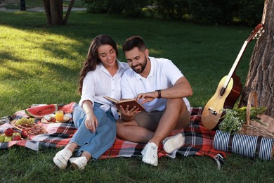 Photo of Happy young couple reading book on plaid in park. Summer picnic