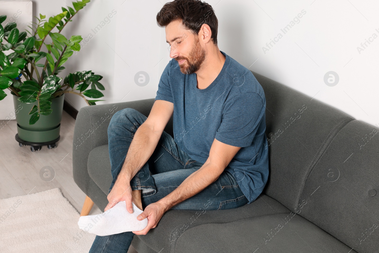 Photo of Man suffering from leg pain and massaging heel on sofa at home