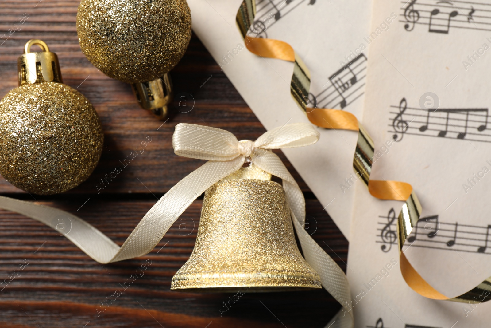 Photo of Golden shiny bell with bow, music sheets and decorative balls on wooden table, flat lay. Christmas decoration