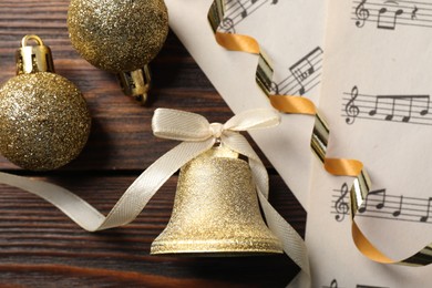 Golden shiny bell with bow, music sheets and decorative balls on wooden table, flat lay. Christmas decoration