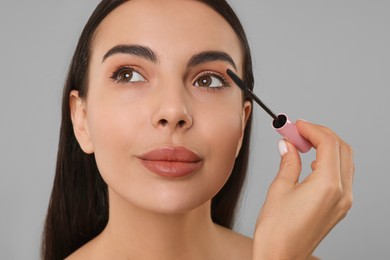 Beautiful young woman applying mascara on grey background, closeup